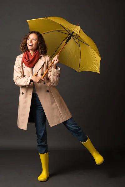 Femme heureuse en trench coat et bottes en caoutchouc posant avec parapluie jaune sur fond noir — Photo de stock