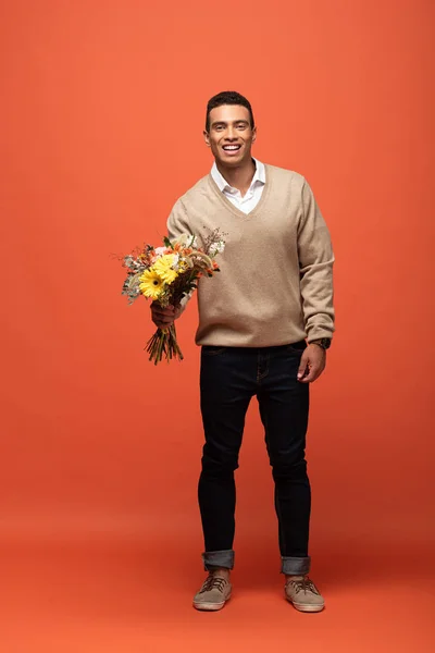 Smiling mixed race man holding autumnal bouquet on orange — Stock Photo