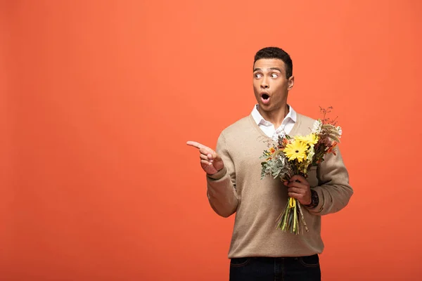 Shocked mixed race man holding autumnal bouquet and pointing with finger aside isolated on orange — Stock Photo