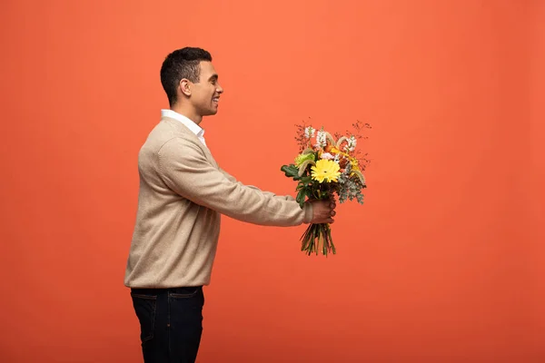 Вид сбоку улыбающегося мужчины смешанной расы, подарившего осенний букет, изолированный на апельсине — стоковое фото