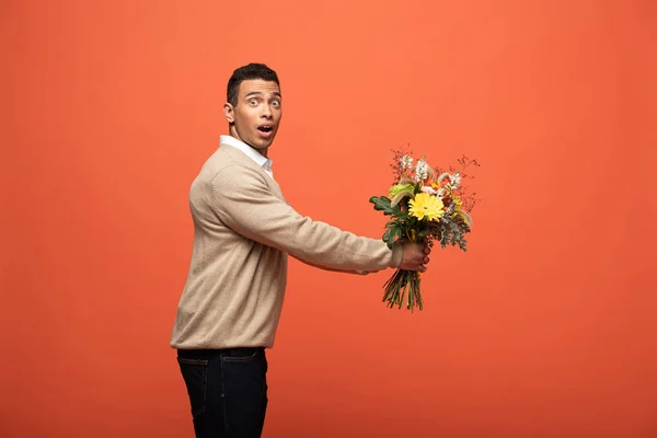 Side view of shocked mixed race man gifting autumnal bouquet isolated on orange — Stock Photo