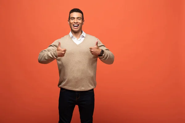 Happy mixed race man in beige sweater showing thumbs up isolated on orange — Stock Photo