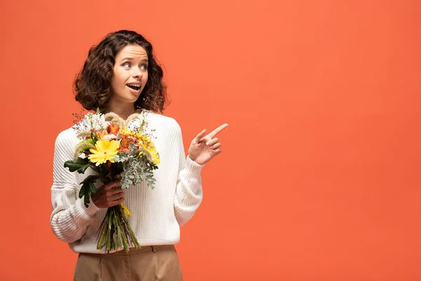 Excited woman in autumnal outfit holding bouquet of flowers and pointing with finger isolated on orange — Stock Photo