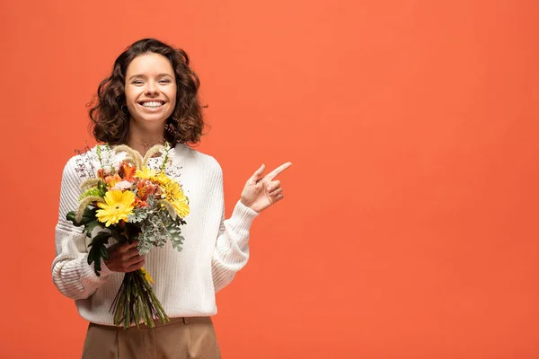 Mujer feliz en traje otoñal sosteniendo ramo de flores y señalando con el dedo aislado en naranja - foto de stock