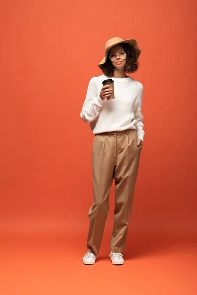 Mujer en sombrero y vasos sosteniendo café para ir en naranja - foto de stock