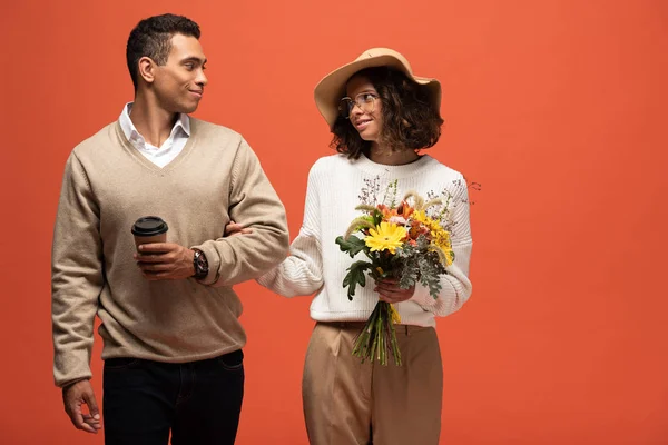 Smiling interracial couple in autumn outfit with coffee to go and bouquet of flowers isolated on orange — Stock Photo