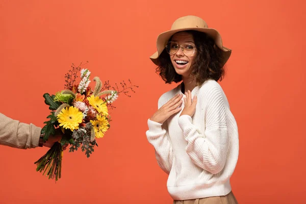 Boyfriend gifting surprised woman in hat bouquet of flowers isolated on orange — Stock Photo