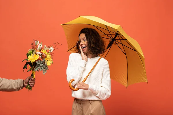 Boyfriend gifting shocked woman with umbrella bouquet of flowers isolated on orange — Stock Photo