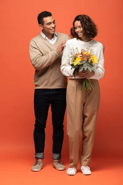 Stylish interracial couple in autumn outfit with bouquet of flowers on orange — Stock Photo