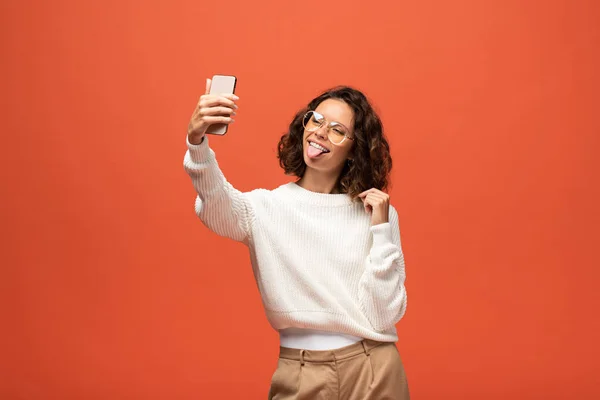 Donna in abito autunnale e occhiali che mostrano la lingua mentre prende selfie isolato su arancione — Foto stock