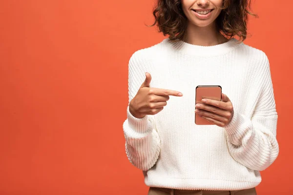 Обрезанный вид улыбающейся женщины в осеннем наряде, указывающей пальцем на смартфон, изолированный на оранжевом — Stock Photo