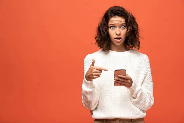 Mujer conmocionada en traje otoñal apuntando con el dedo al teléfono inteligente aislado en naranja - foto de stock