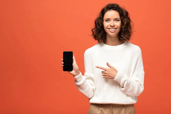 Femme souriante en tenue automnale pointant du doigt le smartphone avec écran blanc isolé sur orange — Photo de stock