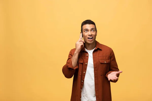 Sorprendido hombre de raza mixta hablando en el teléfono inteligente aislado en amarillo - foto de stock