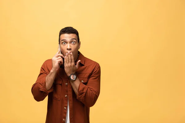 Sorprendido hombre de raza mixta hablando en el teléfono inteligente aislado en amarillo - foto de stock