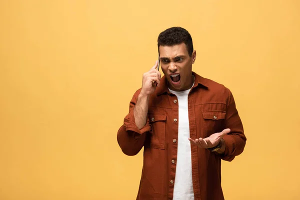 Hombre de raza mixta enojado hablando en el teléfono inteligente y gritando aislado en amarillo - foto de stock