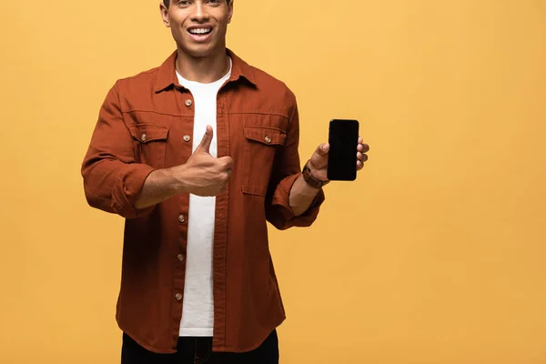 Vue recadrée de sourire mixte homme tenant smartphone avec écran blanc et montrant pouce vers le haut isolé sur jaune — Photo de stock