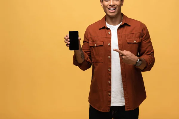 Vista recortada del hombre de raza mixta apuntando con el dedo al teléfono inteligente con pantalla en blanco aislado en amarillo - foto de stock