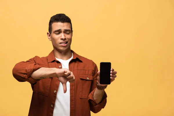 Confused mixed race man showing smartphone with blank screen and thumb down isolated on yellow — Stock Photo