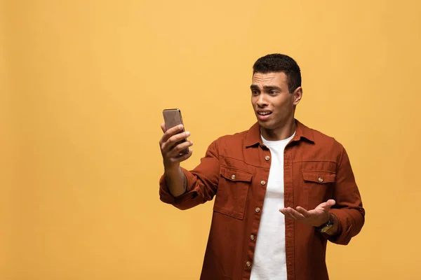 Displeased mixed race man using smartphone isolated on yellow — Stock Photo