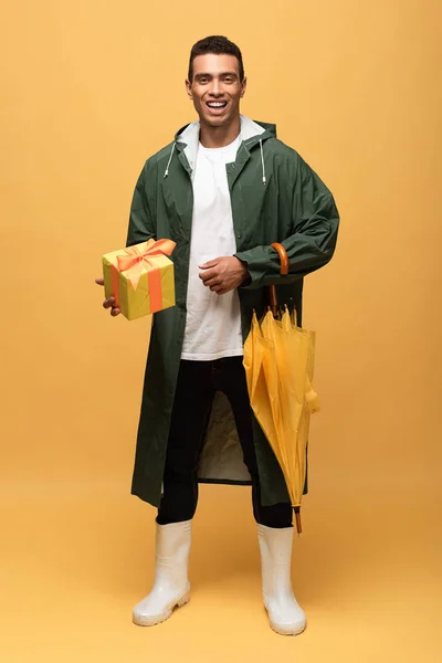 Mixed race man in raincoat and rubber boots holding gift box and umbrella isolated on yellow — Stock Photo