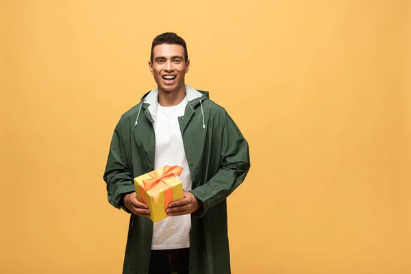 Happy mixed race man in raincoat and rubber boots holding gift box isolated on yellow — Stock Photo