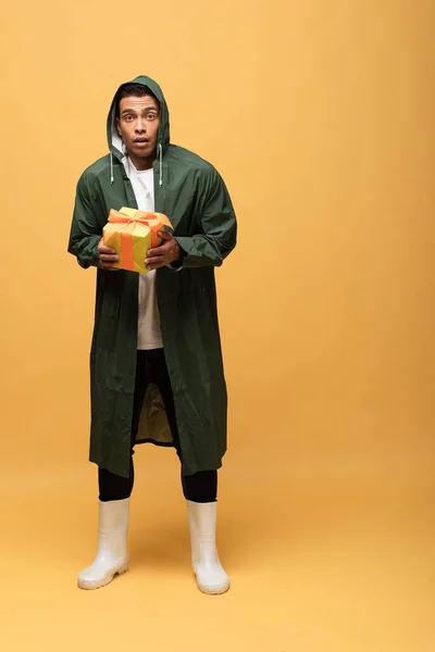 Confused mixed race man in raincoat and rubber boots holding gift box isolated on yellow — Stock Photo