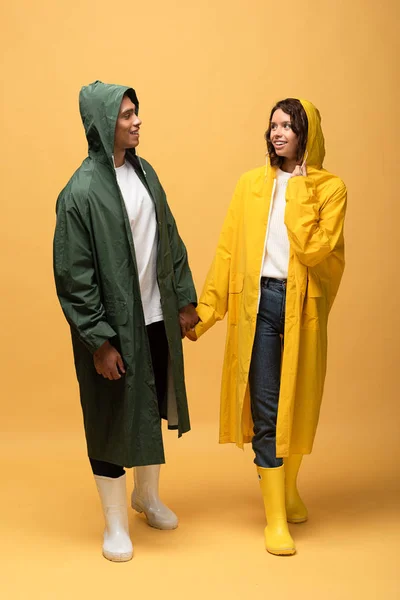 Sonriente pareja interracial en impermeables y botas de goma cogidas de la mano sobre fondo amarillo - foto de stock