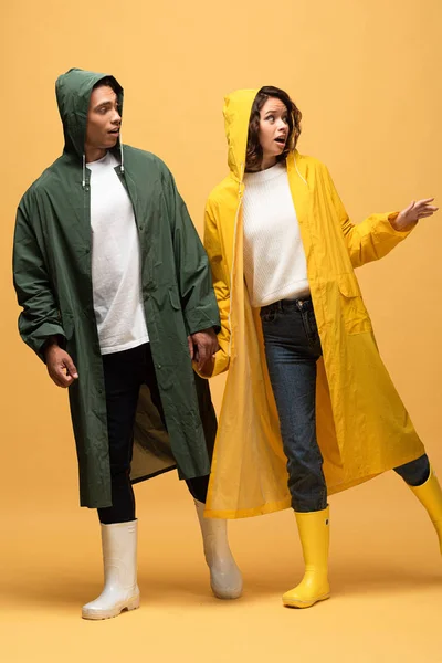 Surprised interracial couple in raincoats and rubber boots on yellow background — Stock Photo