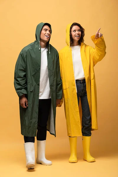 Feliz casal interracial em capas de chuva e botas de borracha apontando com o dedo no fundo amarelo — Fotografia de Stock