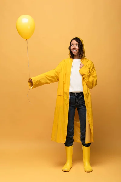 Happy curly woman in yellow raincoat and wellies holding balloon on yellow background — Stock Photo