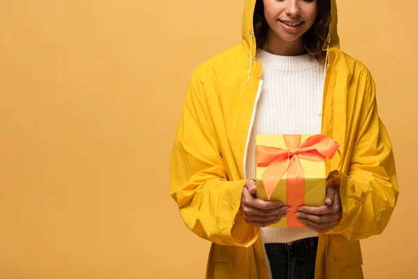 Cropped view of smiling woman in yellow raincoat holding gift box isolated on yellow — Stock Photo