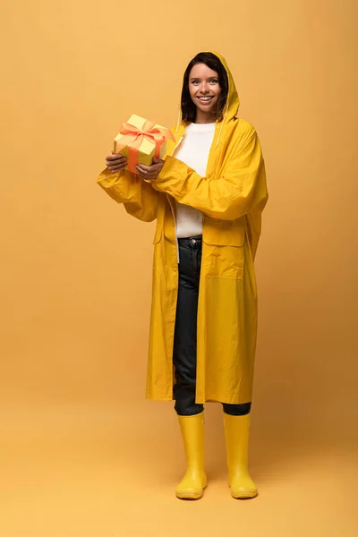 Femme heureuse en imperméable jaune et wellies tenant boîte cadeau sur fond jaune — Photo de stock