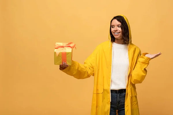 Femme bouclée souriante en imperméable jaune tenant boîte cadeau isolé sur jaune — Photo de stock
