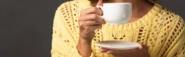 Panoramic shot of woman in yellow knitted sweater holding cup of coffee and saucer on black background — Stock Photo