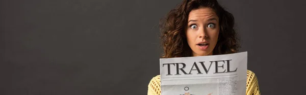 Panoramic shot of shocked curly woman in yellow knitted sweater holding travel newspaper on black background — Stock Photo