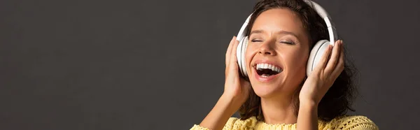 Panoramic shot of happy curly woman with closed eyes in yellow knitted sweater listening music in headphones on black background — Stock Photo