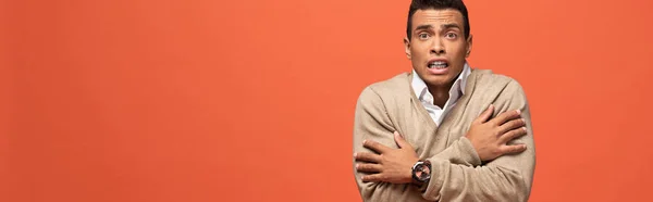 Panoramic shot of confused mixed race man in beige sweater feeling cold isolated on orange — Stock Photo