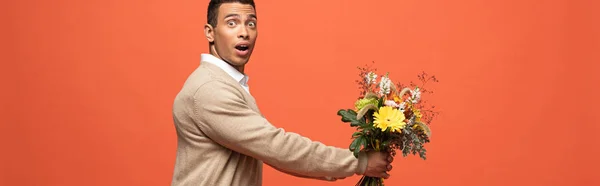 Panoramic shot of shocked mixed race man gifting autumnal bouquet isolated on orange — Stock Photo