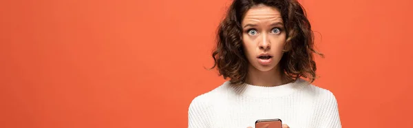 Panoramaaufnahme einer schockierten Frau im herbstlichen Outfit mit Smartphone isoliert auf orange — Stockfoto