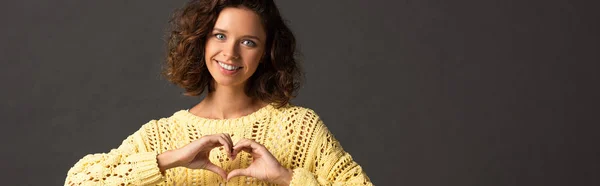 Panoramic shot of happy curly woman in yellow sweater showing heart sign on black background — Stock Photo
