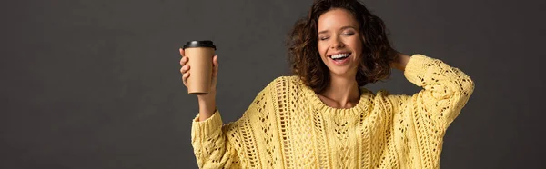Plan panoramique de heureuse femme bouclée en pull tricoté jaune avec les yeux fermés tenant du café pour aller sur fond noir — Photo de stock
