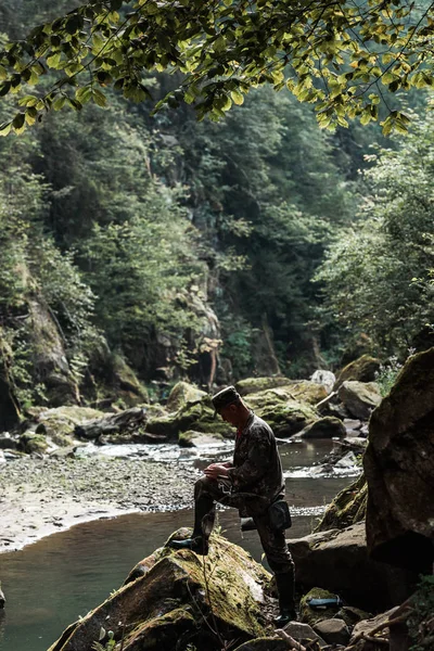 QUIIV, UCRÂNIA - AGOSTO 24, 2019: vista lateral do soldado em uniforme militar perto de água corrente em bosques — Fotografia de Stock