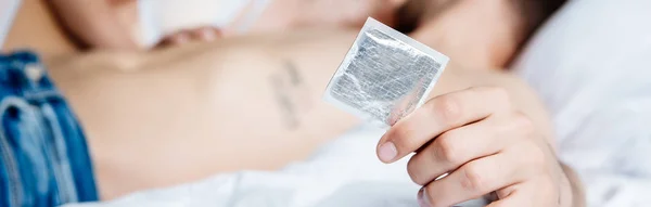 Panoramic shot of man holding condom and lying in bed — Stock Photo
