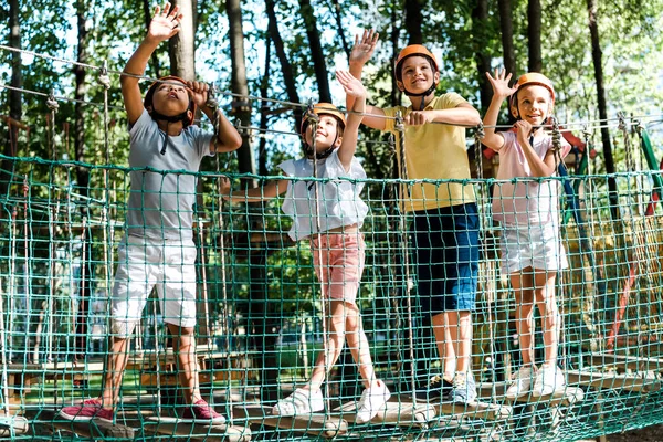 Meninos multiculturais alegres perto de amigos em capacetes acenando as mãos no parque de aventura — Fotografia de Stock