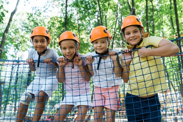 Улыбающиеся дети в оранжевых шлемах рядом с мультикультурными мальчиками в парке приключений — стоковое фото