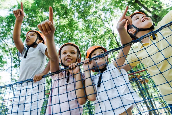 Низкий угол обзора улыбающихся детей в оранжевых шлемах рядом с мультикультурными мальчиками, указывающими пальцами в парке приключений — стоковое фото