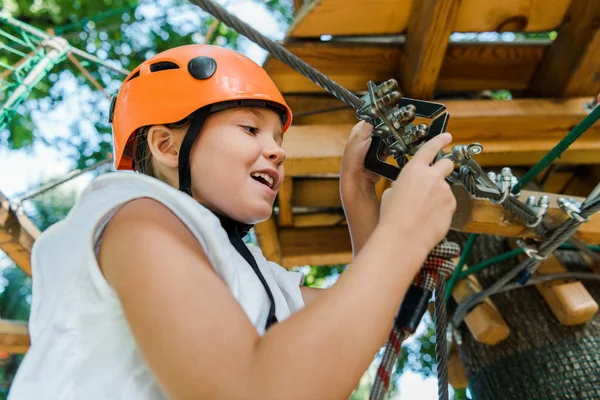 Vista a basso angolo di bambino felice in casco toccare attrezzature di sicurezza nel parco avventura — Foto stock