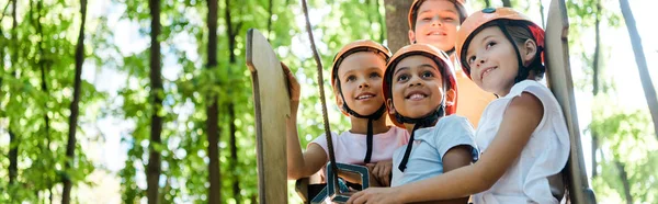 Panoramic shot of positive multicultural kids looking up in adventure park outside — Stock Photo