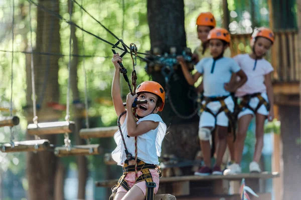 Foco seletivo de criança animado montando em trilha de corda alta perto de crianças multiculturais — Fotografia de Stock